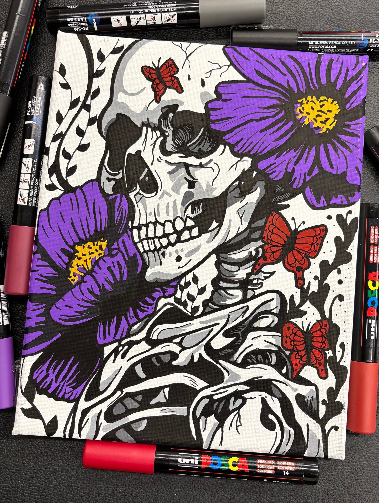 Coloring book with skeleton and flowers design, surrounded by markers on a dark surface.